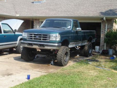1996 Ford F-150 - A lifted green Ford F-Series truck is parked in a driveway next to another truck with its hood open.