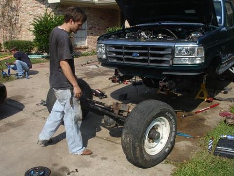 1996 Ford F-150 - A 1996 Ford F-150's front end is disassembled in a driveway, with a person working on the chassis.