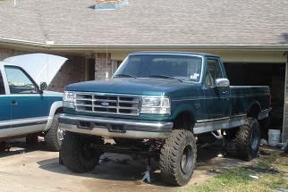 1996 Ford F-150 - A lifted 1996 Ford F-150 pickup truck is parked in front of a garage, with another truck beside it.