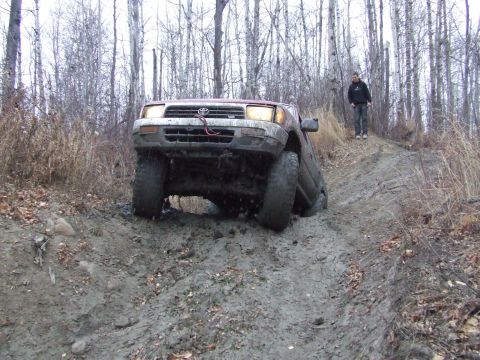 1990 Ford Bronco - A red Toyota 4Runner navigates a muddy incline in a wooded area with a person observing from higher ground.