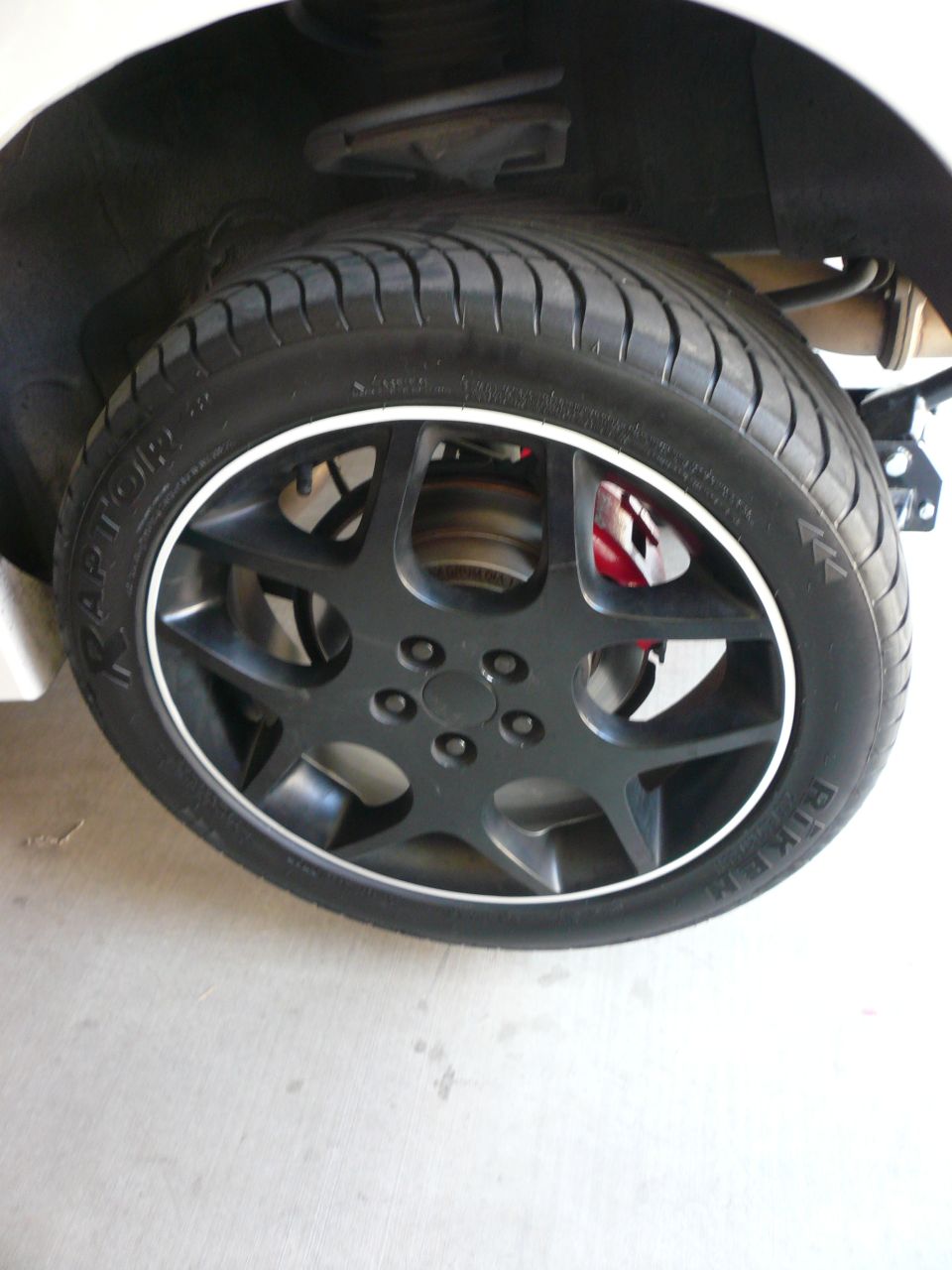 Close-up of a car's black five-spoke wheel with a white trim ring and a Riken Raptor tire. Red brake caliper visible behind the wheel