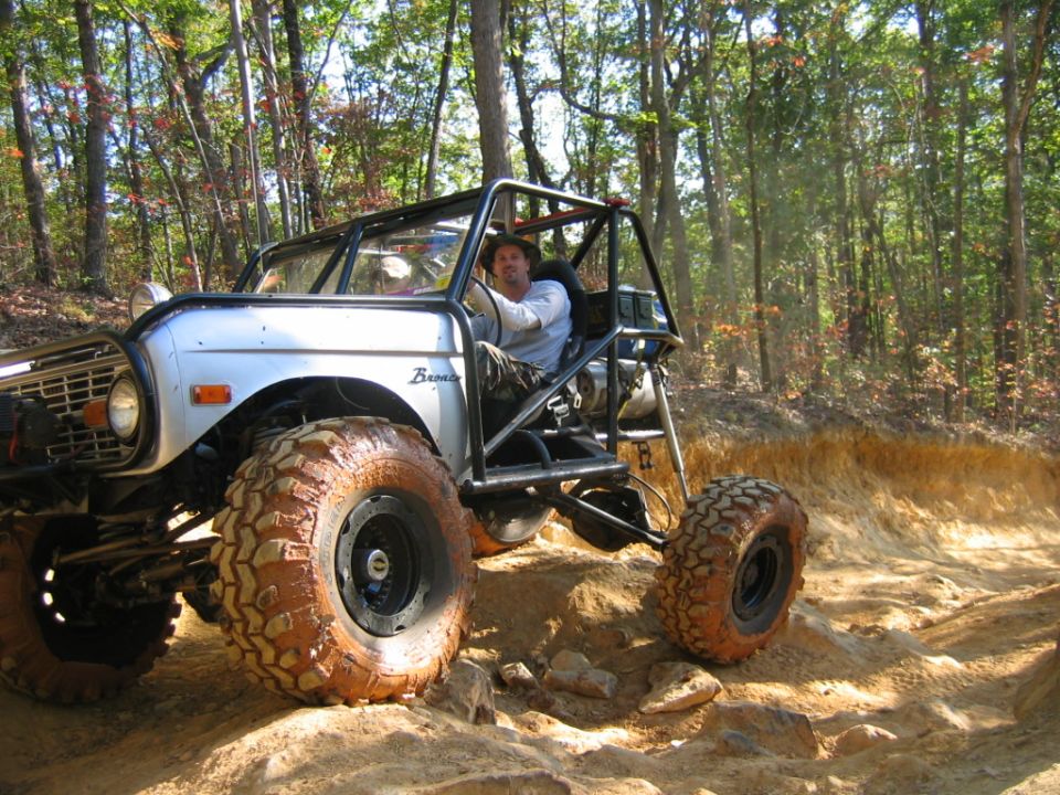 Man driving a modified off-road vehicle with large tires on a dirt trail through the woods