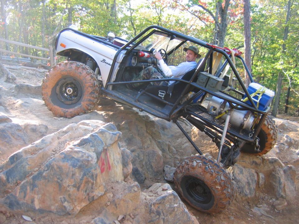 Off-road buggy with a roll cage climbing over rocks. Driver is wearing a hat and camo pants