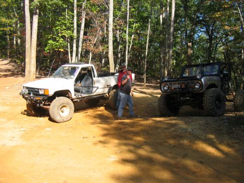 1992 Ford Mustang - Two off-road vehicles are parked on a dirt path surrounded by trees.