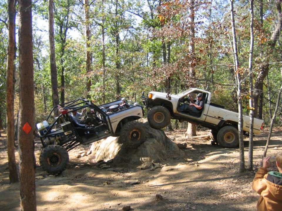 Two off-road trucks navigating a rocky trail in a forest, one climbing over a large rock