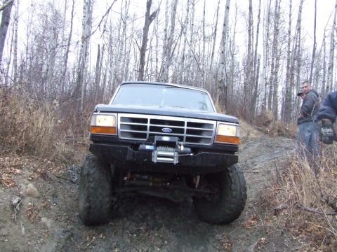 1990 Ford Bronco - A black Ford Bronco is shown from the front, navigating a muddy, wooded terrain.