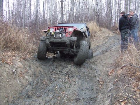 1990 Ford Bronco - A red Jeep Wrangler struggles through thick mud on an off-road trail, with two onlookers standing by.