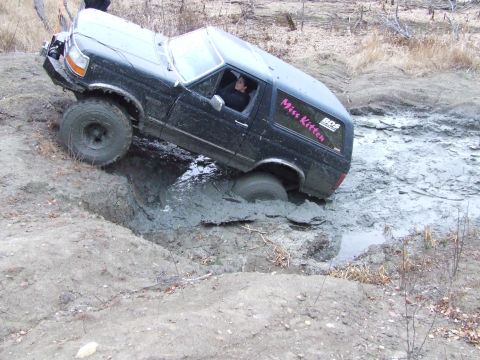 1990 Ford Bronco - A black 1990 Ford Bronco is stuck in a muddy ditch, with its front-right tire deep in the mire.