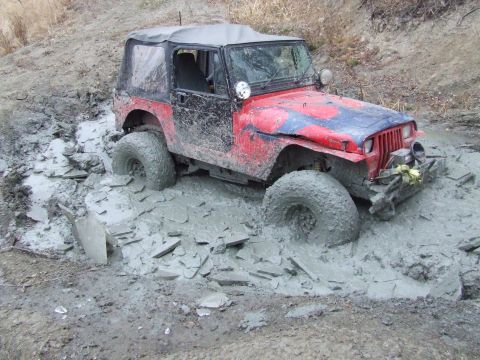 1990 Ford Bronco - A red and blue Jeep Wrangler is stuck in mud, with its front right tire deep in the mire.