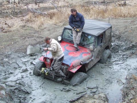 1990 Ford Bronco - Two men attempt to free a red Jeep Wrangler that is deeply stuck in thick mud.