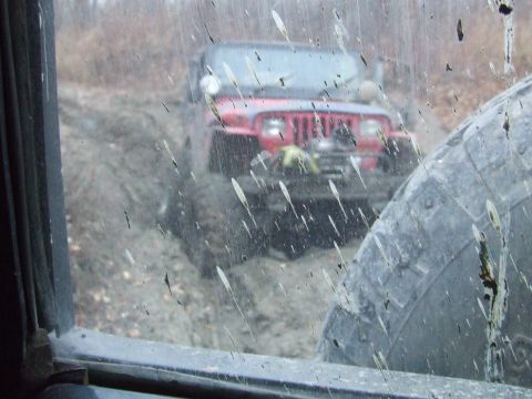 1990 Ford Bronco - A red Jeep Wrangler navigates a muddy trail, viewed through a dirty window.