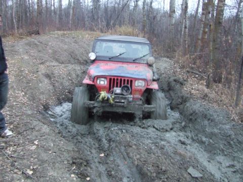 1990 Ford Bronco - A red Jeep Wrangler is stuck in thick mud on a trail surrounded by trees.