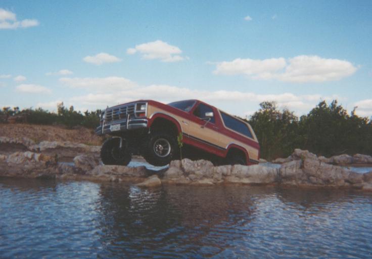 Lifted red and tan Ford Bronco on large tires parked on rocks beside water
