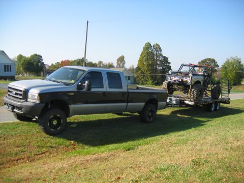 1992 Ford Mustang - A Ford F-250 pickup truck is seen towing a heavily modified off-road vehicle on a trailer under a clear blue sky.