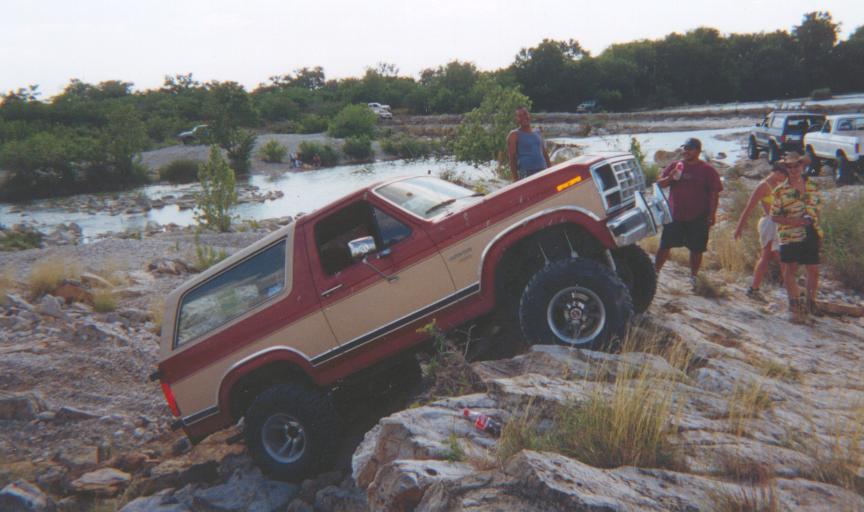 Lifted red and tan Ford Bronco off-roading up rocks by a river as people watch