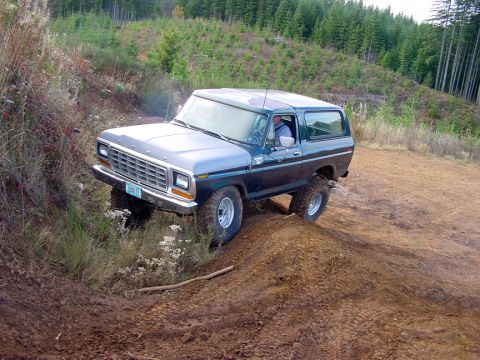 1979 Ford Bronco - A two-tone Ford Bronco navigates an off-road trail, its front left wheel lifted on a mound of dirt.
