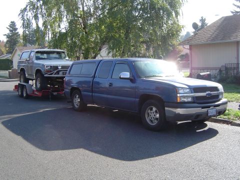 1978 Ford Bronco - A blue pickup truck tows a trailer carrying a silver SUV down a residential street.