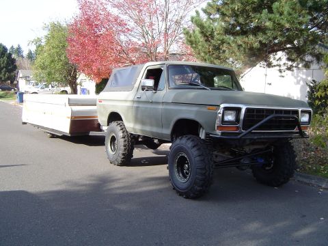 1978 Ford Bronco - Olive green 1978 Ford Bronco with lifted suspension and large tires tows a pop-up camper down a residential street.