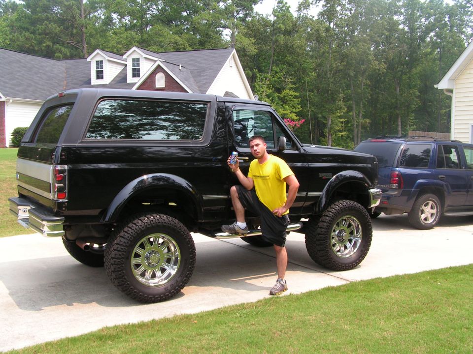 Lifted black Ford Bronco with chrome wheels and big tires; man in yellow shirt posing beside it in a driveway