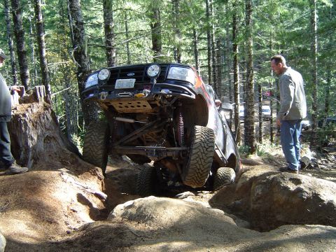 1978 Ford Bronco - An off-road vehicle tackles a challenging rocky obstacle on a wooded trail, with other vehicles visible in the background.