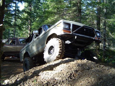 1978 Ford Bronco - Two Ford Broncos off-roading in a wooded area, one climbing over rocks.