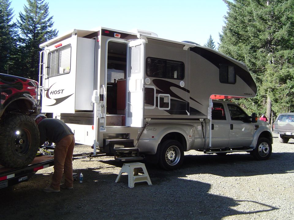 Silver Ford F-series truck with a Host camper shell, parked next to a trailer with a black vehicle. Trees and gravel ground