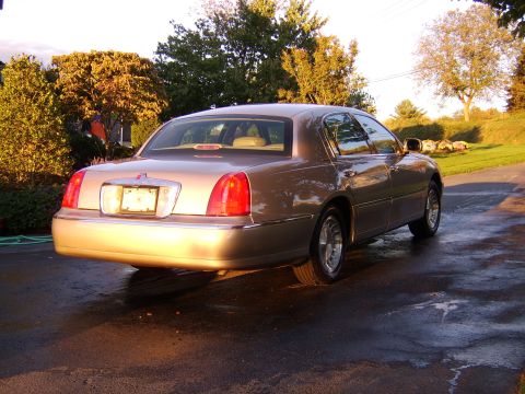 1996 Mercury Grand Marquis - Tan Lincoln Town Car seen from the rear on a wet driveway with trees in the background.