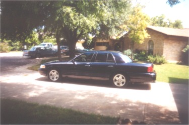 2002 Ford Crown Victoria - A dark sedan is parked in a driveway on a sunny day, with a house and trees in the background.