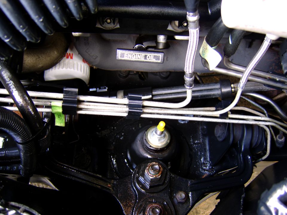 Engine bay close-up with 'ENGINE OIL' cast text, metal fuel lines, braided hoses and yellow-capped fitting