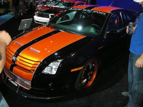 1998 Ford Crown Victoria - black Ford Fusion with orange center stripes sits lowered on orange wheels at a crowded indoor auto show.