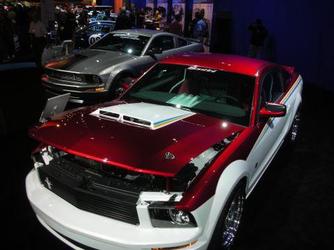 1998 Ford Crown Victoria - red-and-white Ford Mustang with its hood removed sits beside a silver Mustang on a dark show floor.