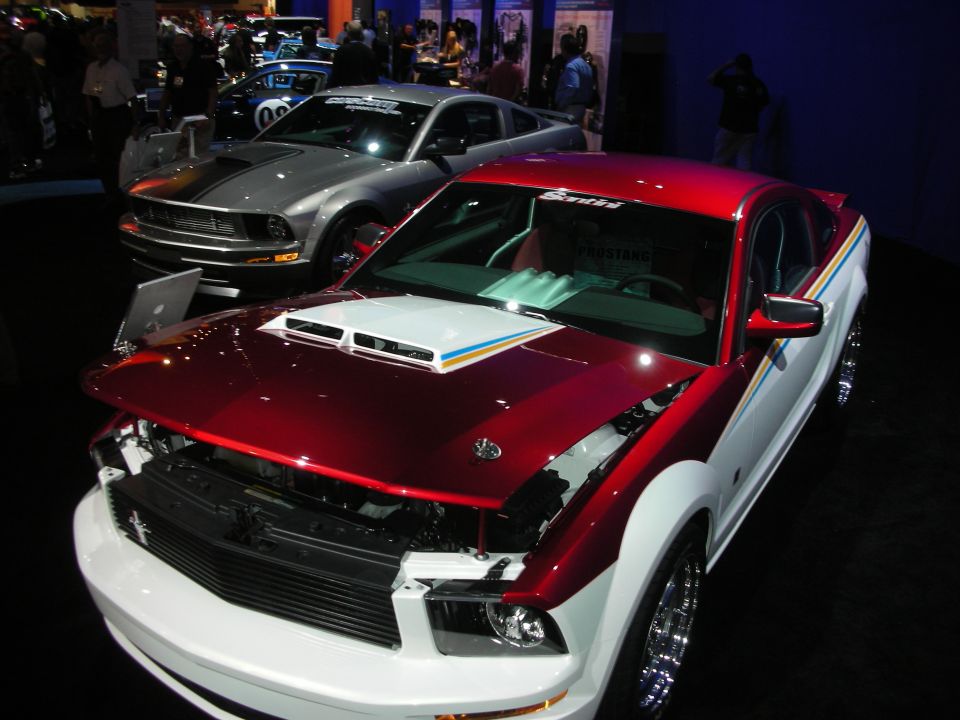 Red and white Ford Mustang with hood off beside silver Mustang at auto show