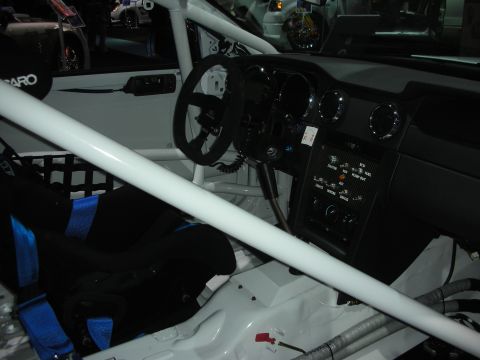 1998 Ford Crown Victoria - Stripped race-car interior showing a white roll cage, black Recaro seat with blue harness and a bare dashboard.