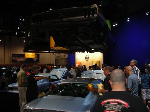 Crowd detailing classic cars at an indoor auto show with a car overhead
