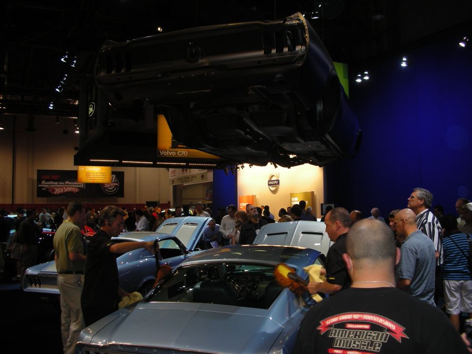 Crowd detailing classic cars at an indoor auto show with a car overhead