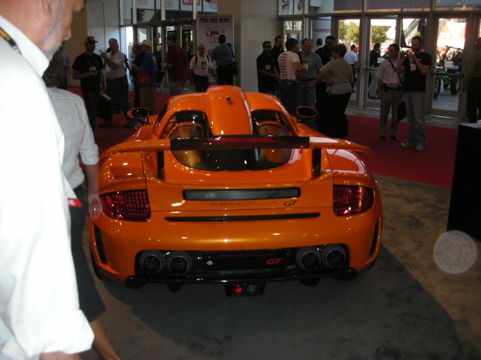Rear view of an orange GT sports car on a show floor near entrance doors