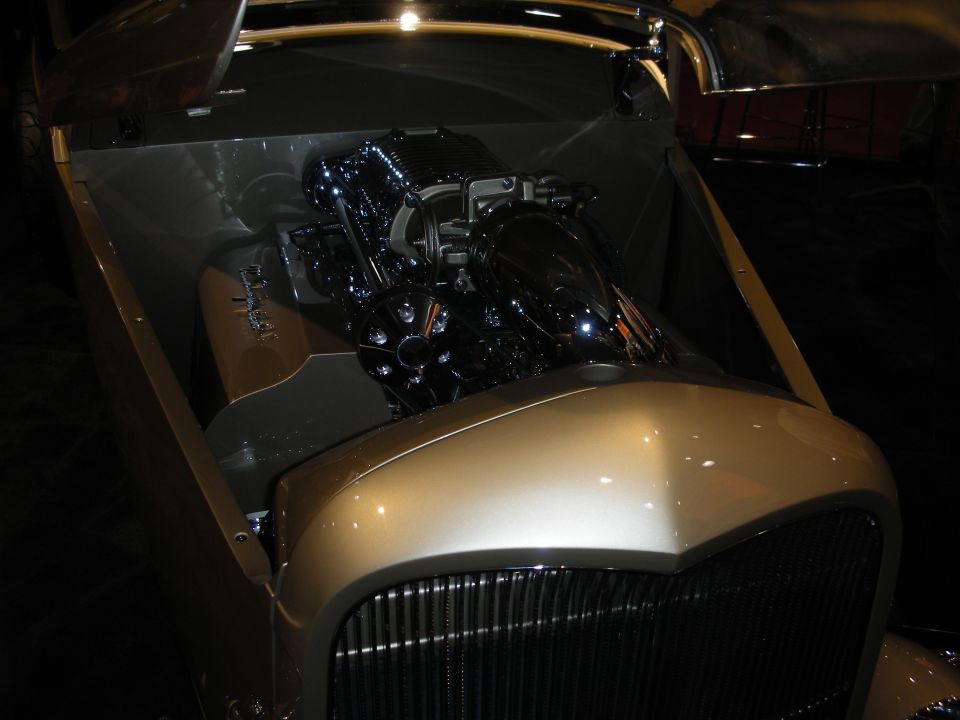 Engine bay of a gold hot rod with a chrome supercharged V8 and vertical grille