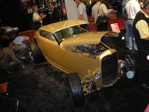 Gold open-wheel hot rod roadster on display at an indoor car show with spectators