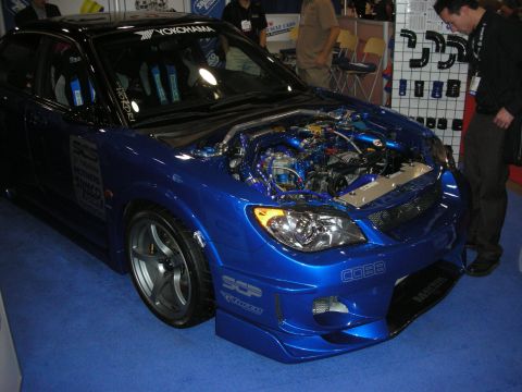 Blue tuner sedan with hood open showing engine at a Yokohama-branded show booth