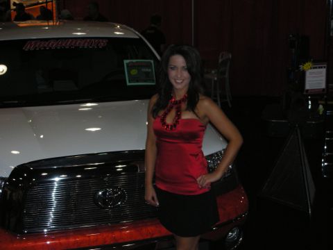 Woman in red top and black skirt posing beside a white pickup truck at a show
