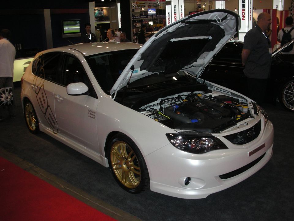 White hatchback with open hood and gold wheels displayed at an indoor car show