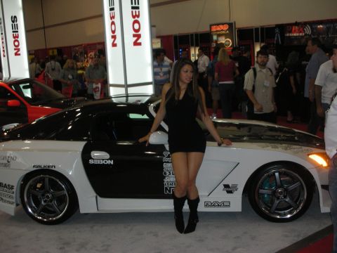 Woman in black dress posing beside a white wide-body coupe at a Seibon show booth