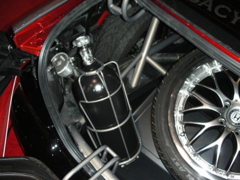 Caged black fire-suppression bottle beside spare tire and silver mesh wheel
