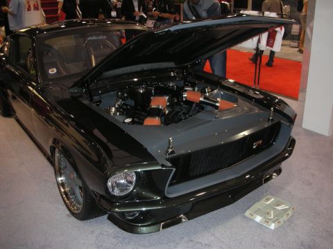 1998 Ford Crown Victoria - black fastback muscle car displayed with its hood raised, revealing a custom engine bay, on a show floor with red carpet.
