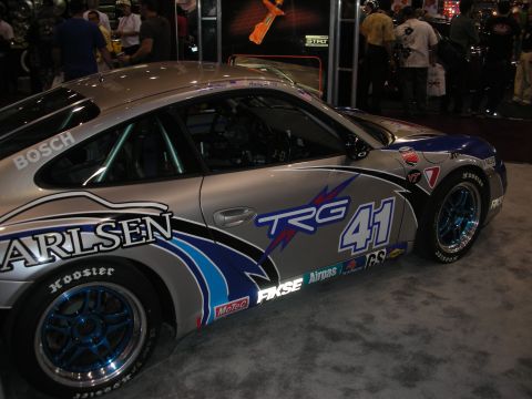 1998 Ford Crown Victoria - silver Porsche race car wearing number 41 and TRG and Carlsen sponsor livery with blue wheels, displayed at an indoor show.