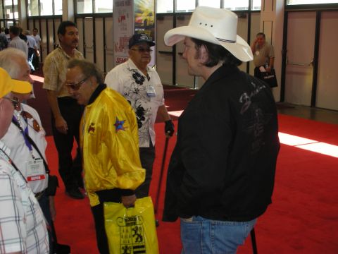 1998 Ford Crown Victoria - man in a white cowboy hat converses with an older man wearing a yellow star-emblem jacket near a red-carpeted show entrance.