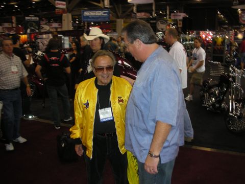 1998 Ford Crown Victoria - older man in a yellow star jacket smiles while standing with a man in a blue shirt on a busy indoor show floor.