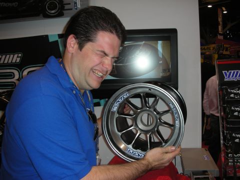 1998 Ford Crown Victoria - man in a blue polo grins while holding up a polished RAYS-branded racing wheel at a trade show booth.