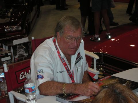 1998 Ford Crown Victoria - older man wearing an Edelbrock-branded shirt and lanyard signs an autograph at a table during an indoor show.