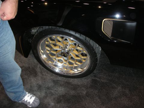 1998 Ford Crown Victoria - black custom car wheel with a chrome and gold mesh design and low-profile tire, beside a person in jeans on show carpet.